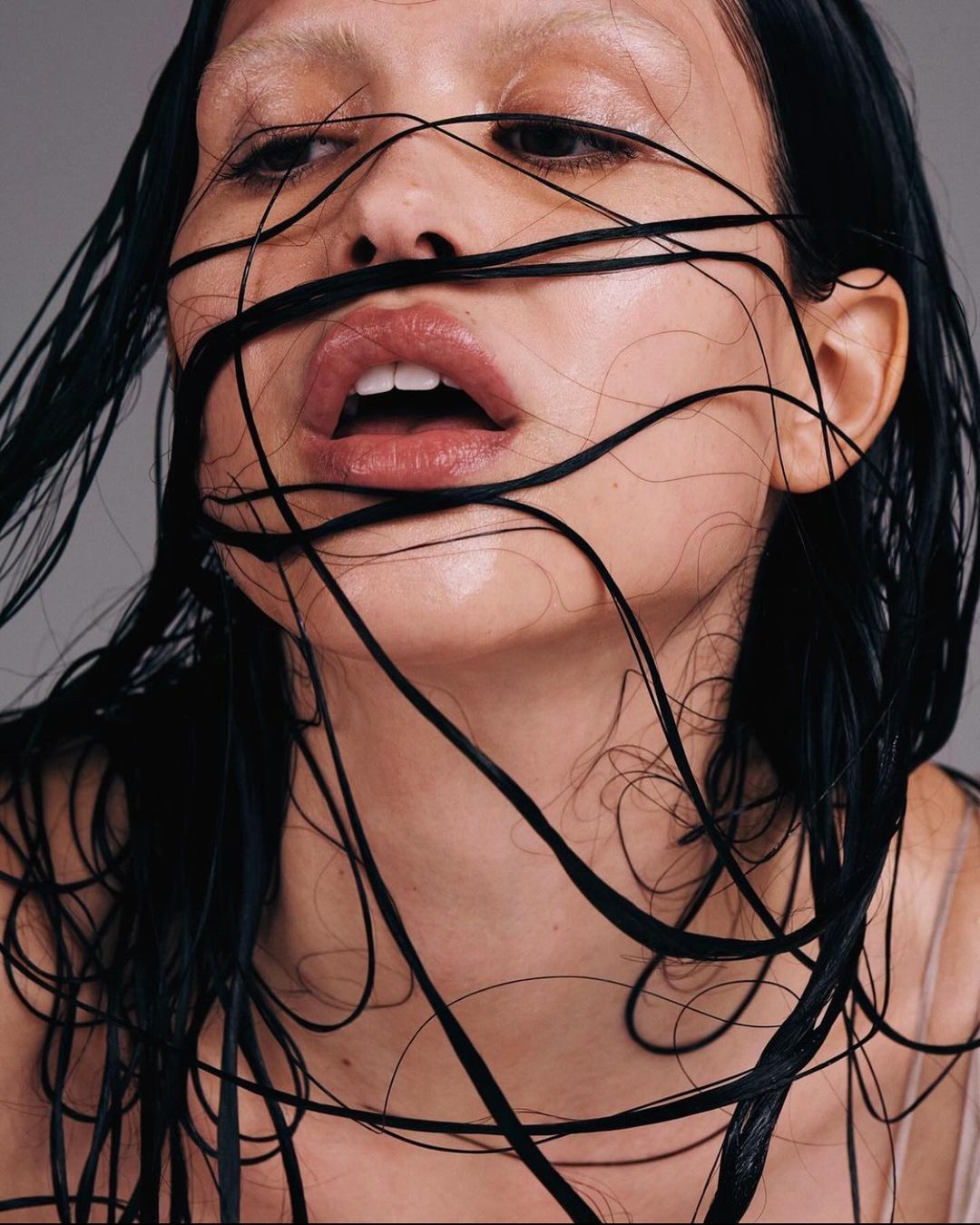 Close-up of a woman's face with wet dark hair draped across her face, pink lips slightly parted, pale skin and light brows.