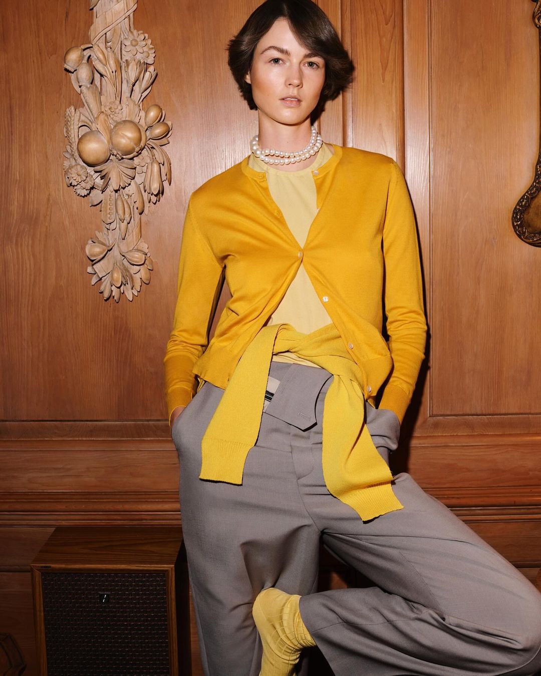 A fashion shot of a woman in gray slacks, mustard cardigan, pearl necklace and yellow socks posing indoors against a wood paneled wall.