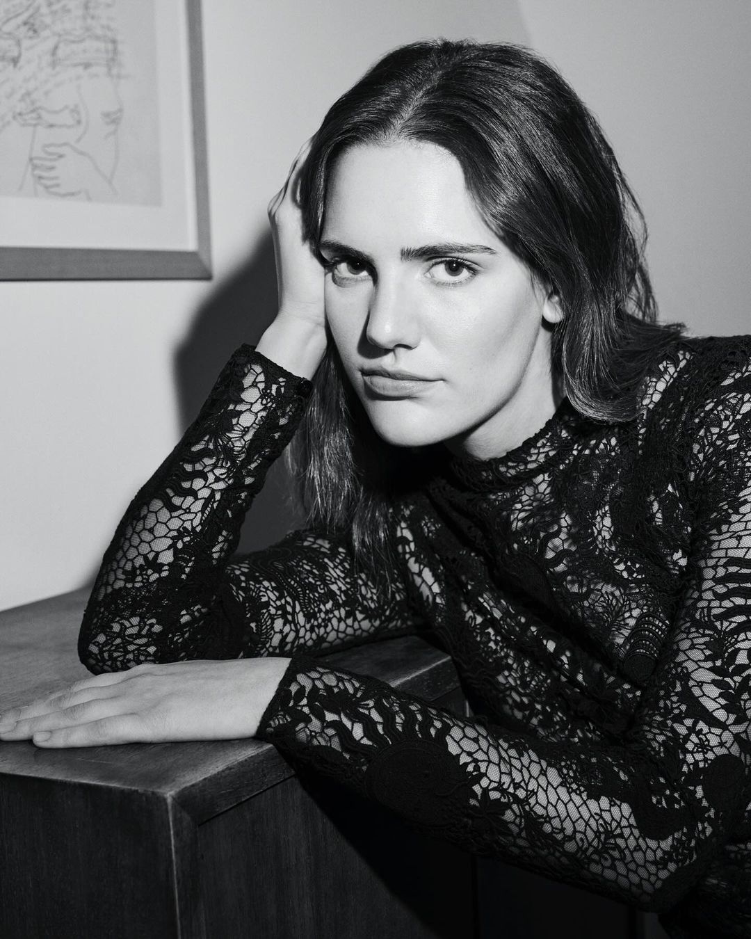 Portrait of an adult woman in a black lace dress resting her head on her hand, posing near a dark wood cabinet in a black and white photo.