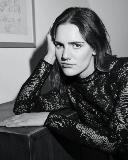 Portrait of an adult woman in a black lace dress resting her head on her hand, posing near a dark wood cabinet in a black and white photo.