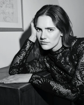 Portrait of an adult woman in a black lace dress resting her head on her hand, posing near a dark wood cabinet in a black and white photo.