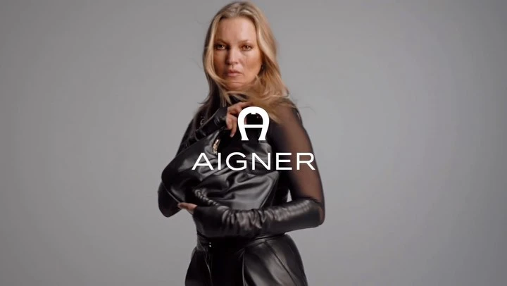 Kate Moss poses with an AIGNER black leather handbag, wearing a black top and skirt against a neutral grey studio background.