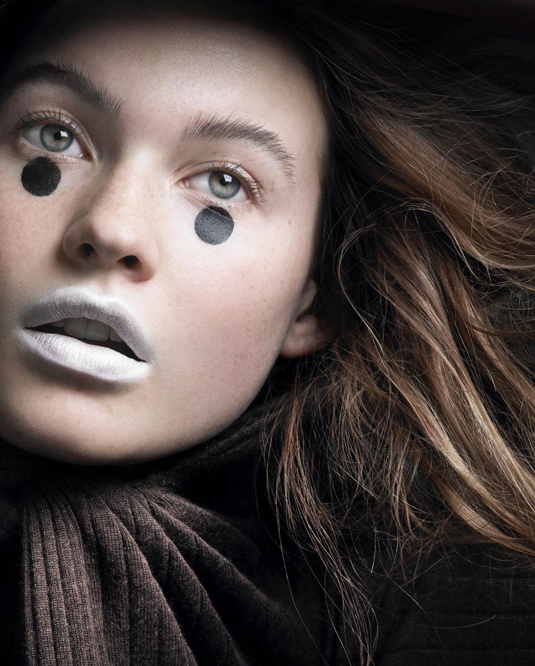 Portrait of a young woman with brown hair and white lipstick, featuring black dots as makeup under her eyes, posing indoors.