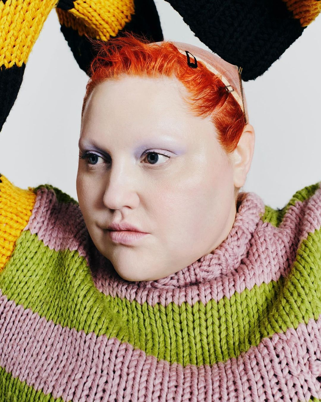Close-up of a redhead woman in a bright, chunky striped knit sweater, posing against a neutral background in a studio setting.
