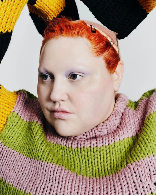 Close-up of a redhead woman in a bright, chunky striped knit sweater, posing against a neutral background in a studio setting.