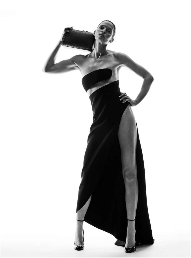Black-and-white studio shot of an adult female fashion model in a black dress with a high slit and heels, carrying a clutch.