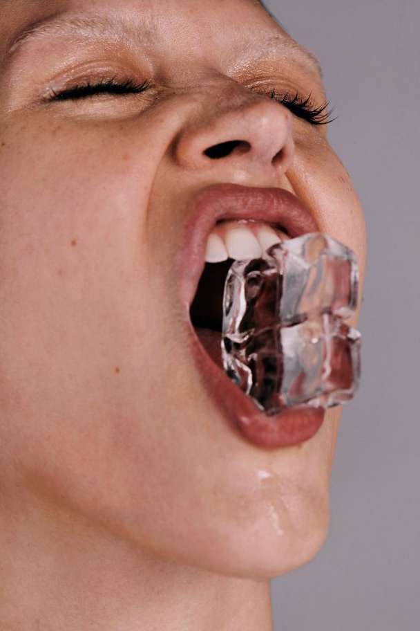 Close-up portrait of a young fair-skinned person with eyes closed and an ice cube in their mouth, against a light gray background.