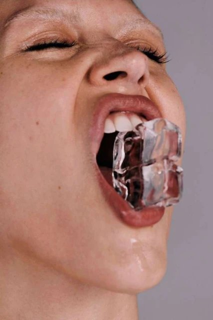 Close-up portrait of a young fair-skinned person with eyes closed and an ice cube in their mouth, against a light gray background.