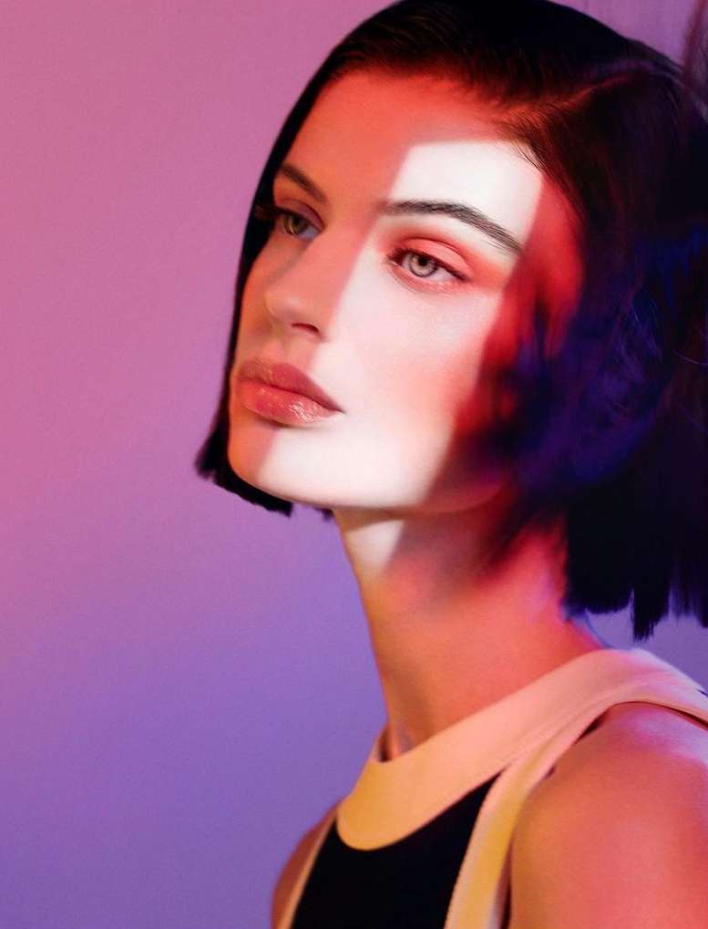 Portrait of a young adult female model with auburn-toned makeup, in a black-and-white top, posing in front of a red and purple background.