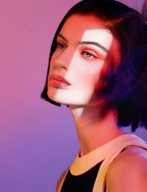 Portrait of a young adult female model with auburn-toned makeup, in a black-and-white top, posing in front of a red and purple background.