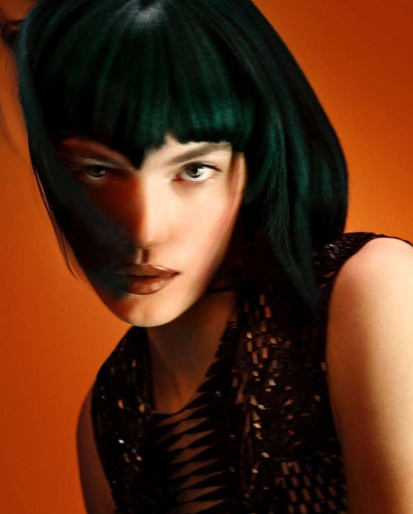 Close-up portrait of a woman with dark green hair against a solid orange background, showcasing beauty and fashion elements.