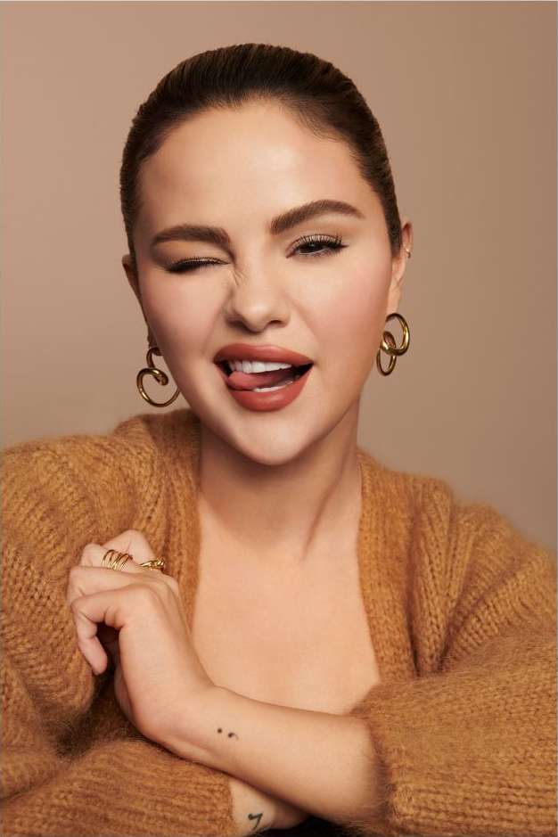 Selena Gomez winking and sticking out her tongue, wearing a fuzzy brown cardigan and gold hoop earrings against a beige backdrop.