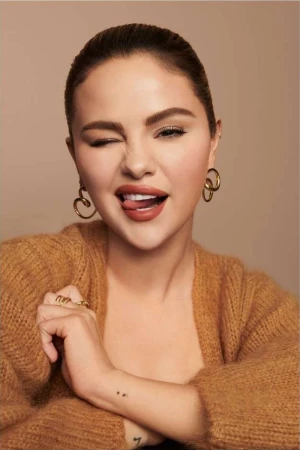 Selena Gomez winking and sticking out her tongue, wearing a fuzzy brown cardigan and gold hoop earrings against a beige backdrop.