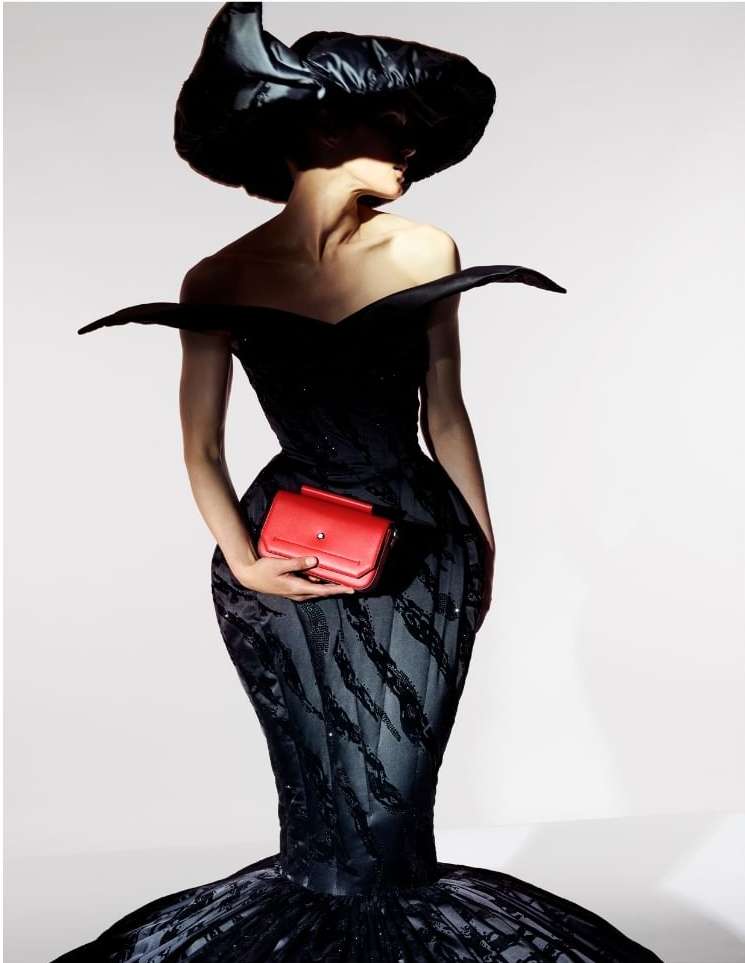 Adult female fashion model wears a black couture gown with a black hat and holds a red clutch bag in a studio setting.
