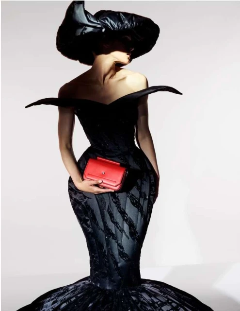 Adult female fashion model wears a black couture gown with a black hat and holds a red clutch bag in a studio setting.