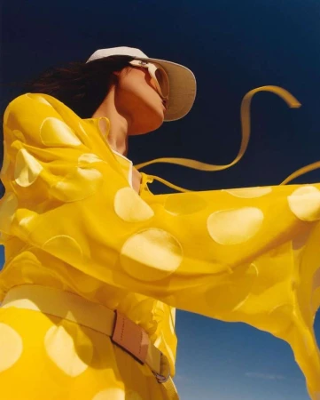 Model in a yellow polka dot dress and white cap looking up against a dark blue sky, photographed by Liz Collins.