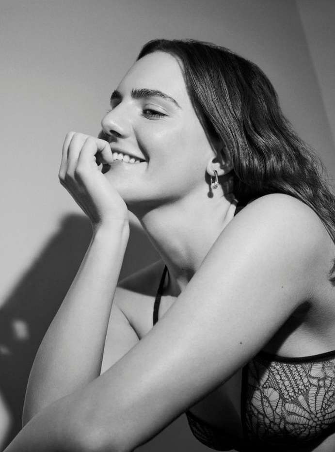 Black-and-white portrait of a smiling young woman with long hair, wearing a lace top and earrings, resting her chin on her hand.