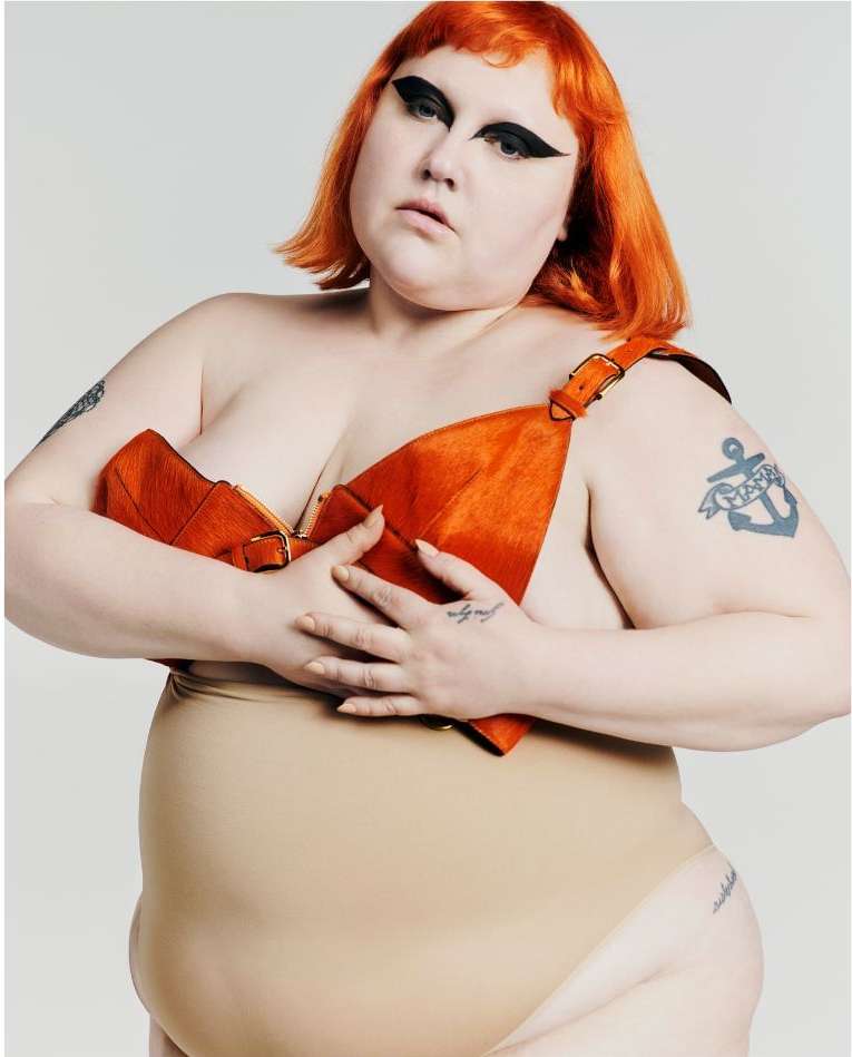 Plus-size woman with bright orange hair and creative makeup in an eye-level portrait in a studio against a white background.