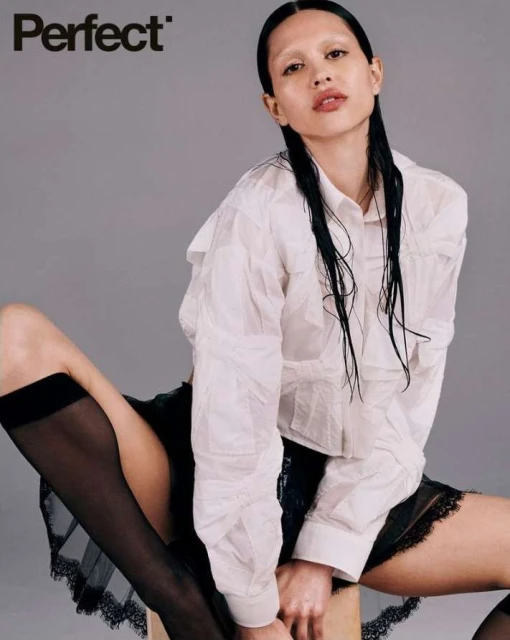 Portrait of a woman with damp hair, wearing a white shirt and black skirt with sheer black thigh-high stockings, posing against a gray background.