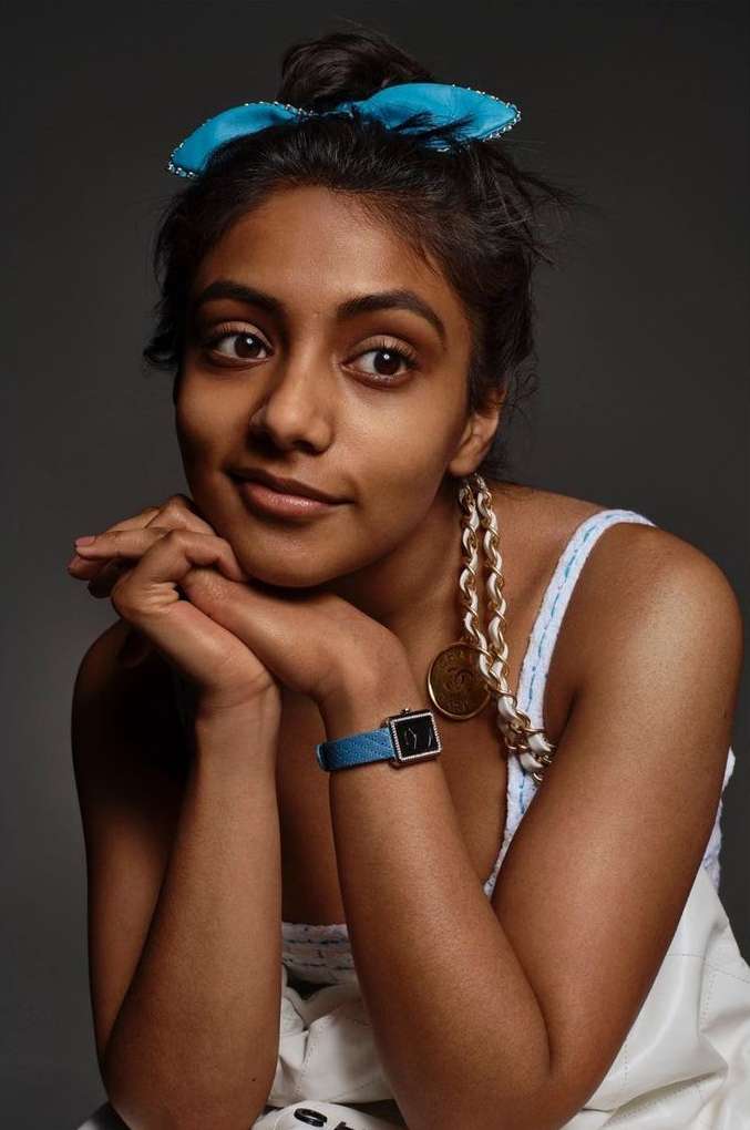 Portrait of a young woman with a blue hair bow and large gold necklace, resting her chin on her hands in a studio setting.