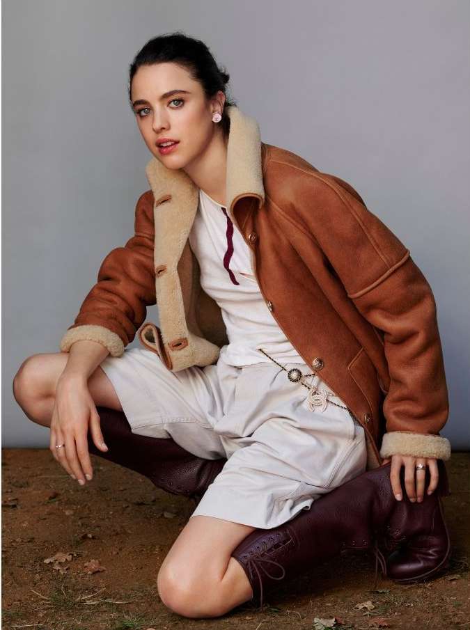 Portrait of a young adult woman kneeling outdoors, wearing a brown shearling jacket, white shorts, and riding boots, with a grey background.