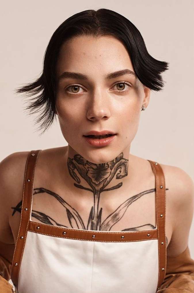 Close-up portrait of a woman with short black hair and a floral tattoo on her chest and neck, wearing a white top with brown straps.