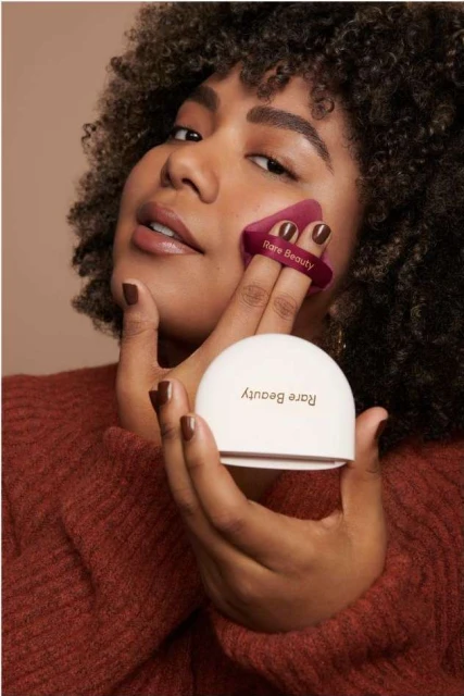 Woman with curly hair applies Rare Beauty makeup with a cosmetic sponge in a close-up shot against a beige backdrop.