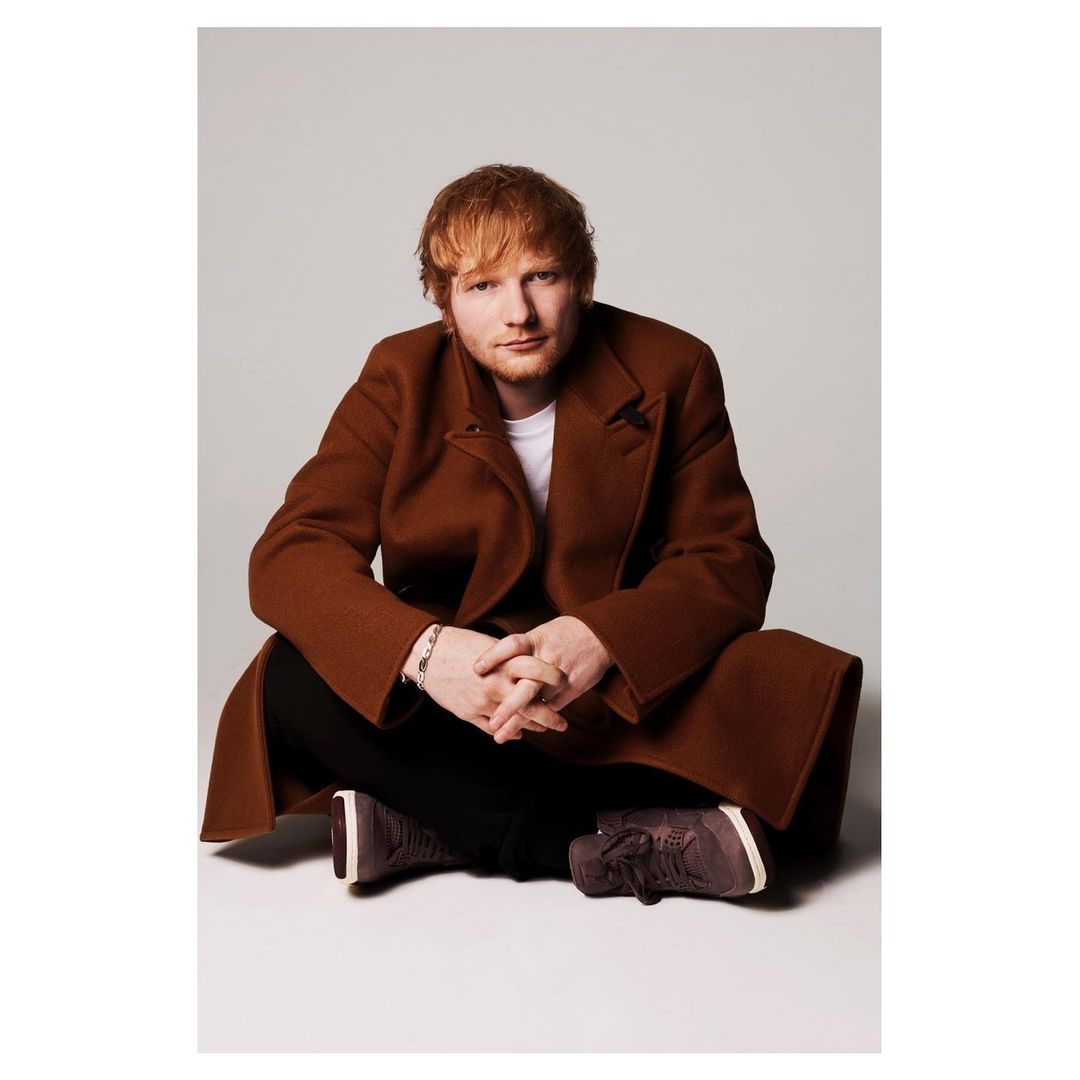 Adult male, Ed Sheeran, in studio portrait wearing brown coat and black pants sitting cross-legged on white background looking at the camera.