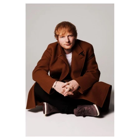 Adult male, Ed Sheeran, in studio portrait wearing brown coat and black pants sitting cross-legged on white background looking at the camera.