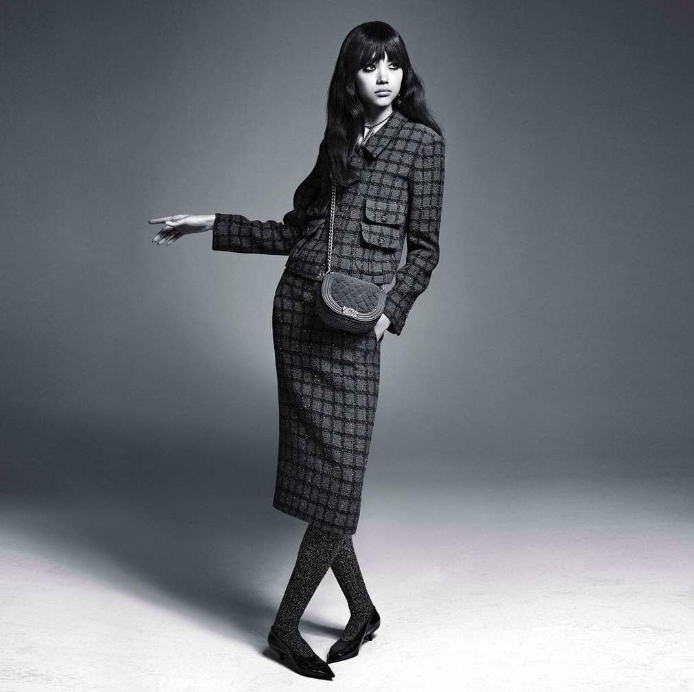 Woman in patterned suit and sparkly tights poses against a grey backdrop with a small bag. Fashion editorial image with a vintage aesthetic.