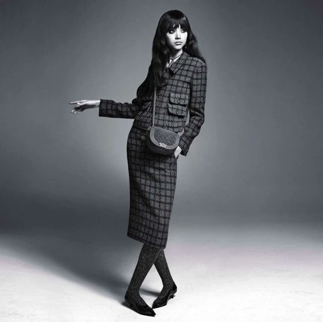 Woman in patterned suit and sparkly tights poses against a grey backdrop with a small bag. Fashion editorial image with a vintage aesthetic.