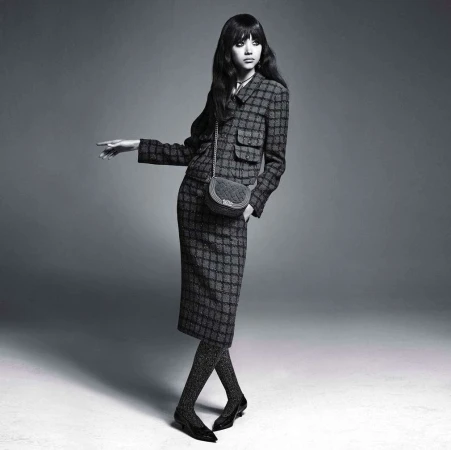 Woman in patterned suit and sparkly tights poses against a grey backdrop with a small bag. Fashion editorial image with a vintage aesthetic.