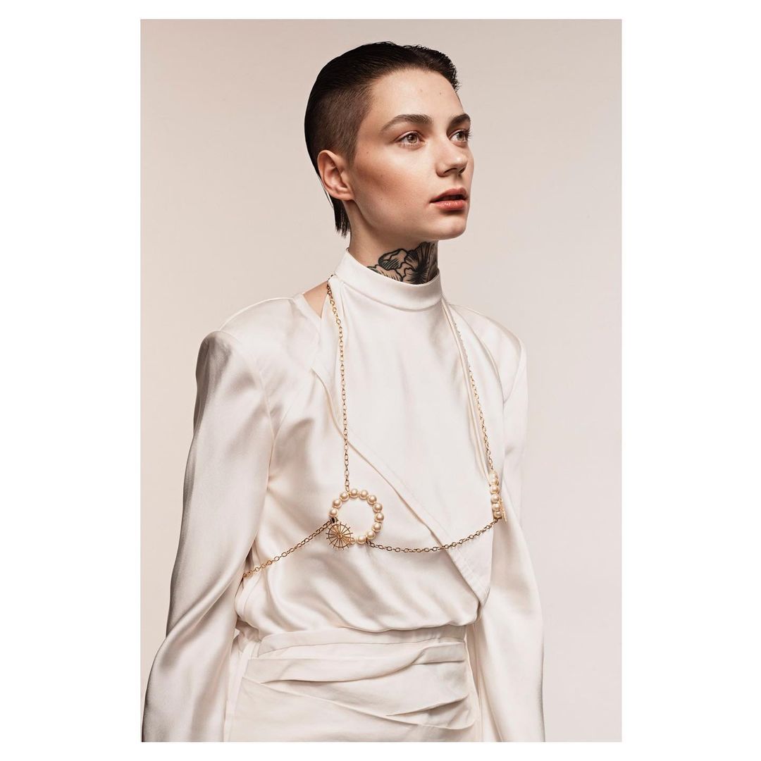Elegant portrait of a young woman in a white dress and gold jewelry, looking off camera with short hair and visible neck tattoo.