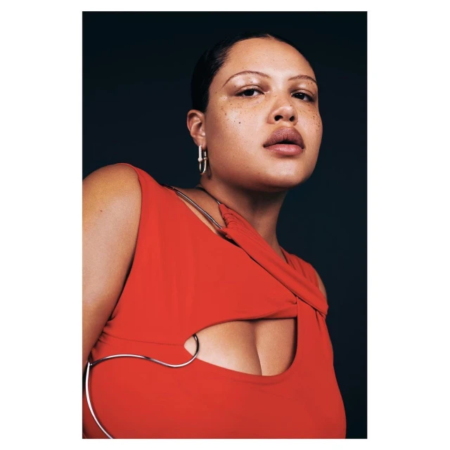 Portrait of a woman with bare shoulders wearing an orange top and hoop earrings, set against a dark blue background in a photo studio.