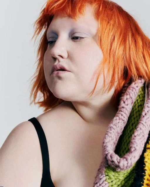 Portrait of a plus-size woman with orange hair and blue eyeshadow wearing a black tank top and multicolor knitted scarf.