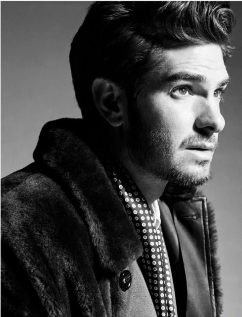 Black and white portrait of an adult male wearing a fur coat and a patterned scarf looking off-camera against a neutral background.