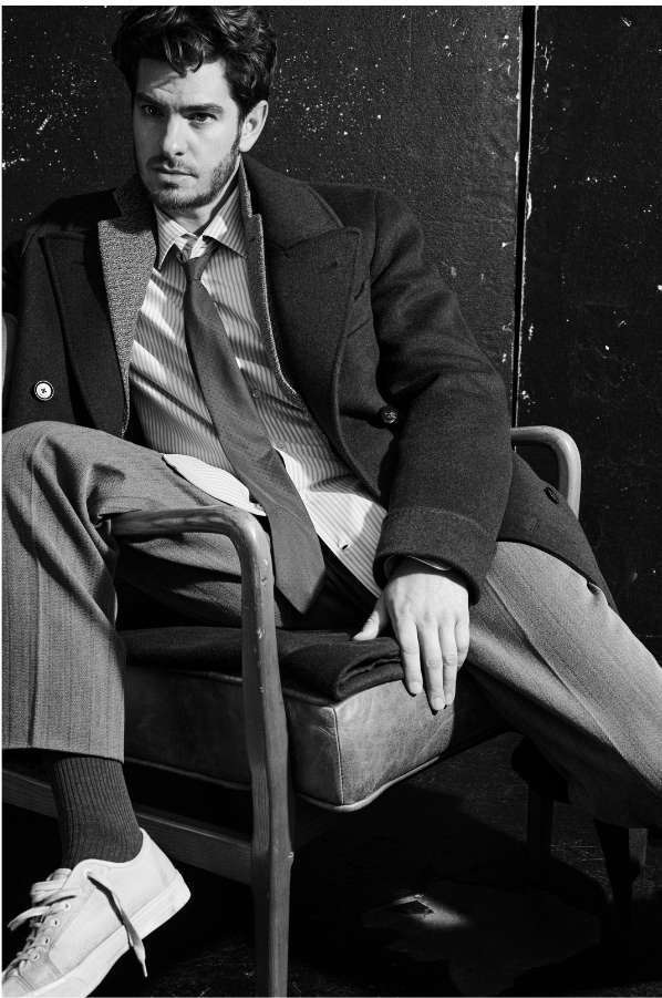 Monochrome portrait of Andrew Garfield seated in an armchair wearing a tailored dark coat, striped shirt, tie, and casual sneakers.