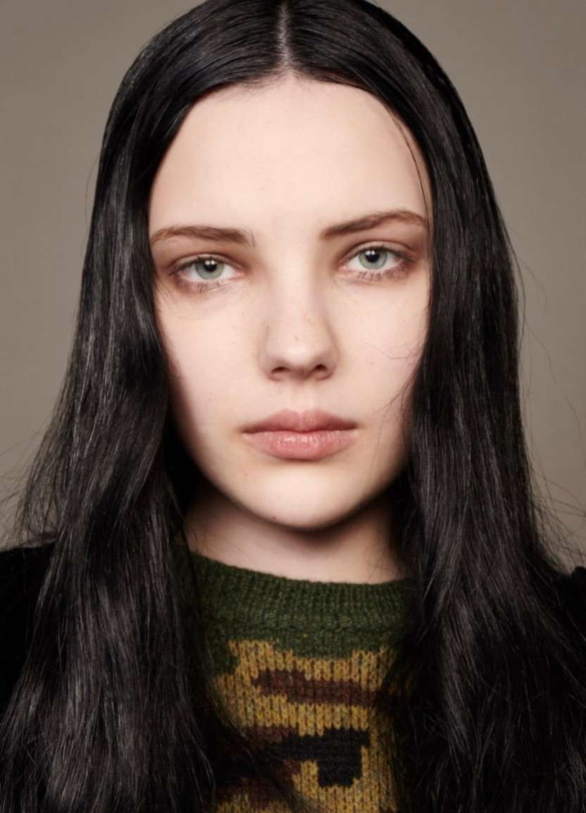 Close-up portrait of a young adult female with fair skin, long black hair, and a patterned sweater, set against a neutral background.