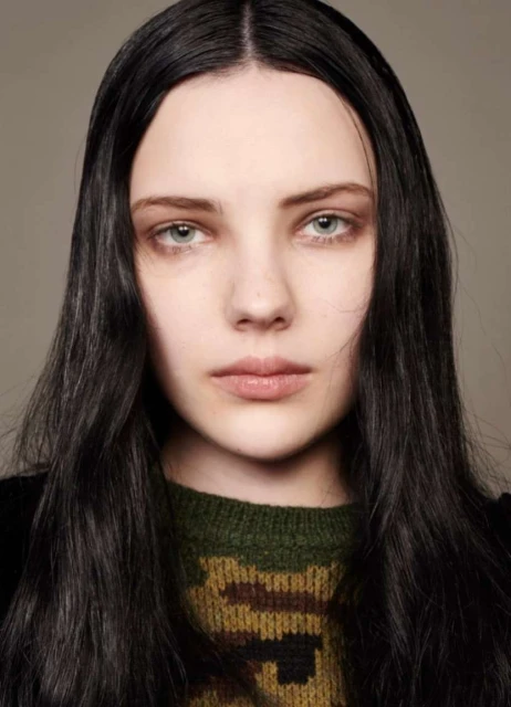 Close-up portrait of a young adult female with fair skin, long black hair, and a patterned sweater, set against a neutral background.
