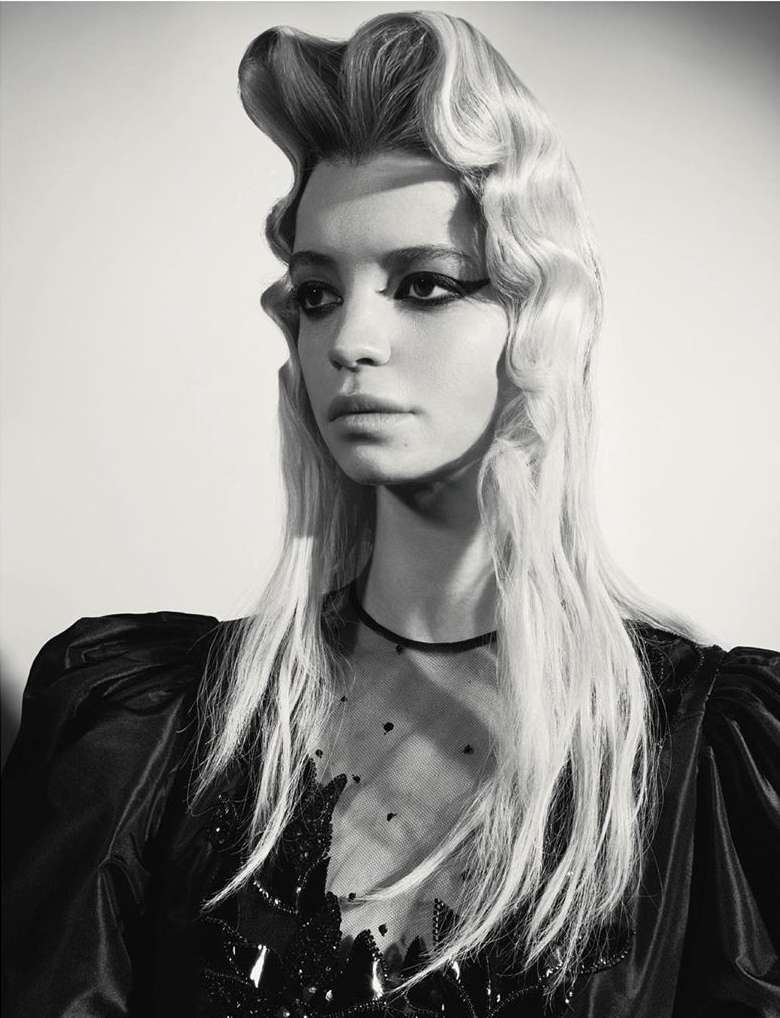 Monochrome portrait of a woman with dramatic eye makeup and a waved hairstyle, posing in a sheer-sleeved dress against a plain background.