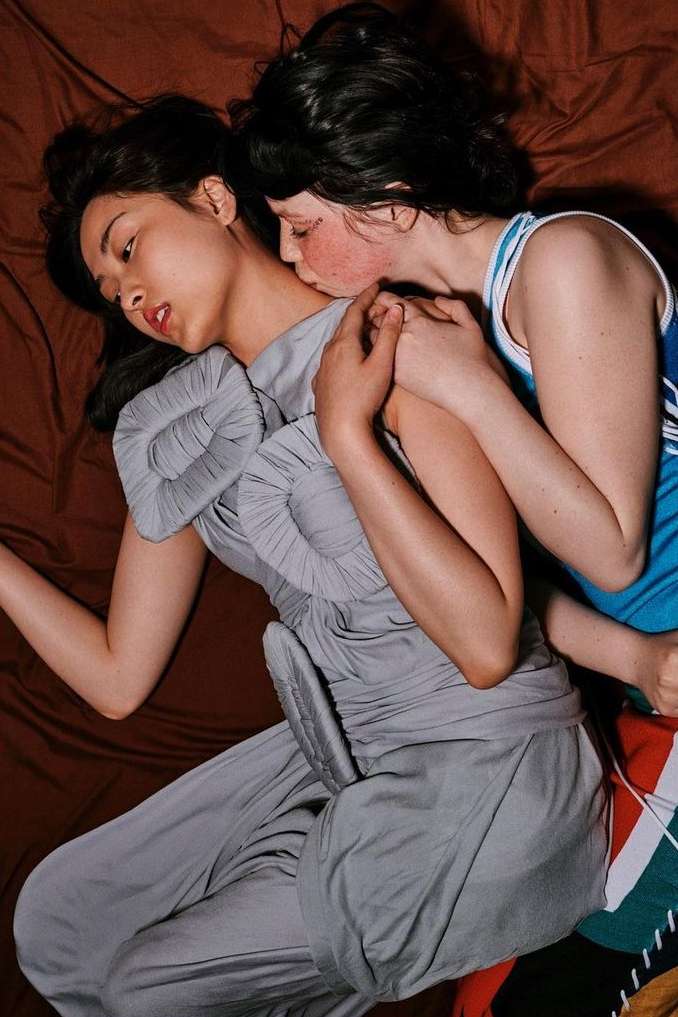 Two women embrace on brown bedding. One woman in a grey dress is lying down while the other in a tank top kisses her neck.