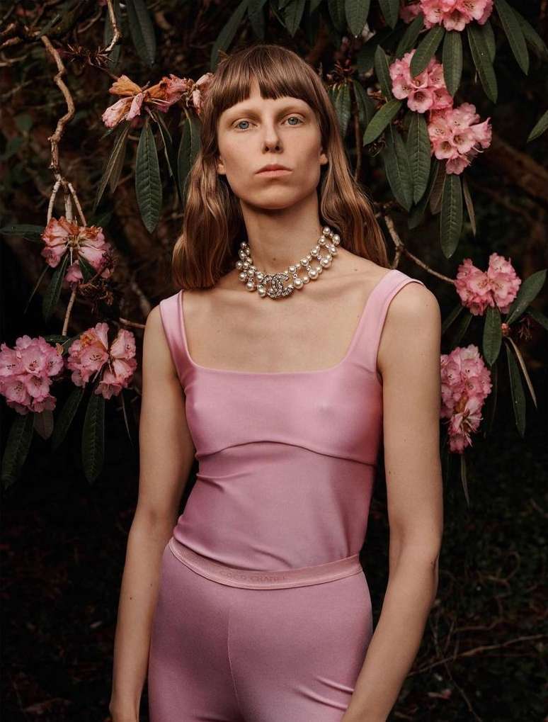 Woman in pink outfit poses in front of blooming pink rhododendrons with a pearl necklace in a medium portrait shot.