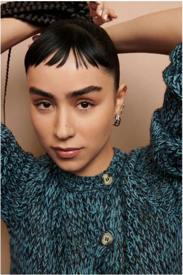 Close-up shot of a young woman with dark hair and blue sweater looking directly at the camera in a studio setting with neutral background.