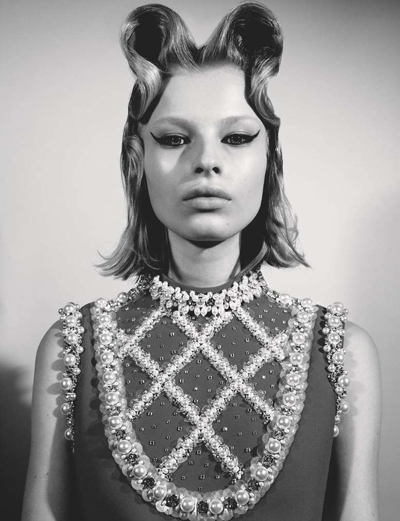 Portrait of a young woman with sculptural retro hairstyle, dramatic eyeliner, and jeweled embellishments in a studio setting.