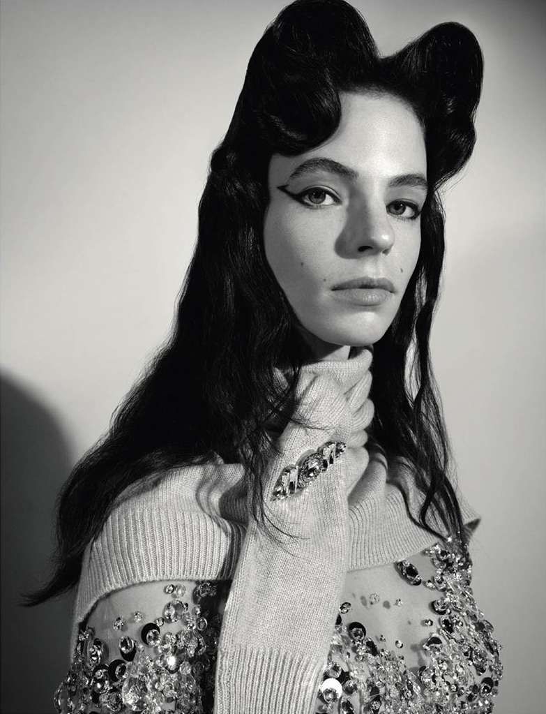 Black and white portrait of a woman with stylized hair and winged eyeliner, wearing a sheer, embellished top and a knit turtleneck.