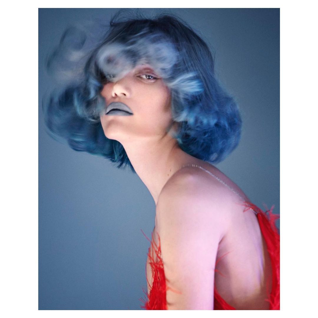 Close-up portrait of adult female model with blue hair and dramatic make-up, posing in front of blue background with blurred smoky effect.