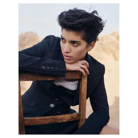 Model with dark hair wearing a black blazer resting chin on hands over a wooden chair outdoors