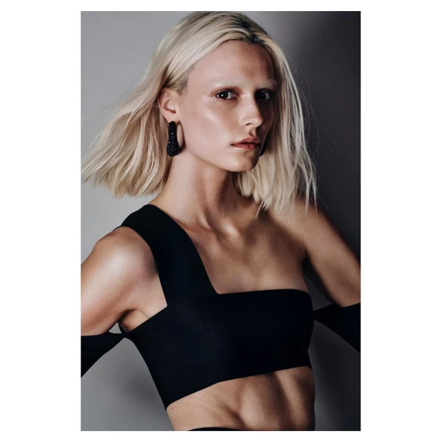 Portrait of a fair-skinned woman with platinum blonde bob haircut, wearing a black one-shoulder top, and earrings against a grey background.