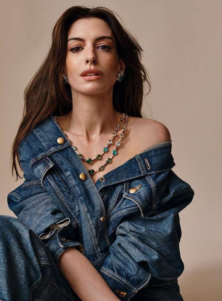 Anne Hathaway poses wearing a denim jacket and jeans with a sparkling necklace in a studio shot with a neutral background.