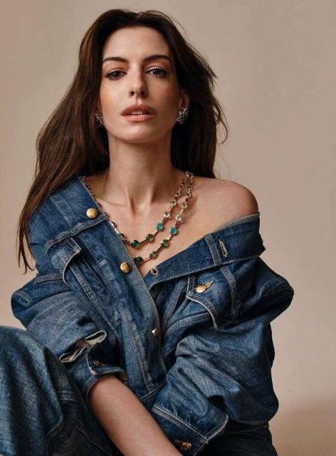 Anne Hathaway poses wearing a denim jacket and jeans with a sparkling necklace in a studio shot with a neutral background.